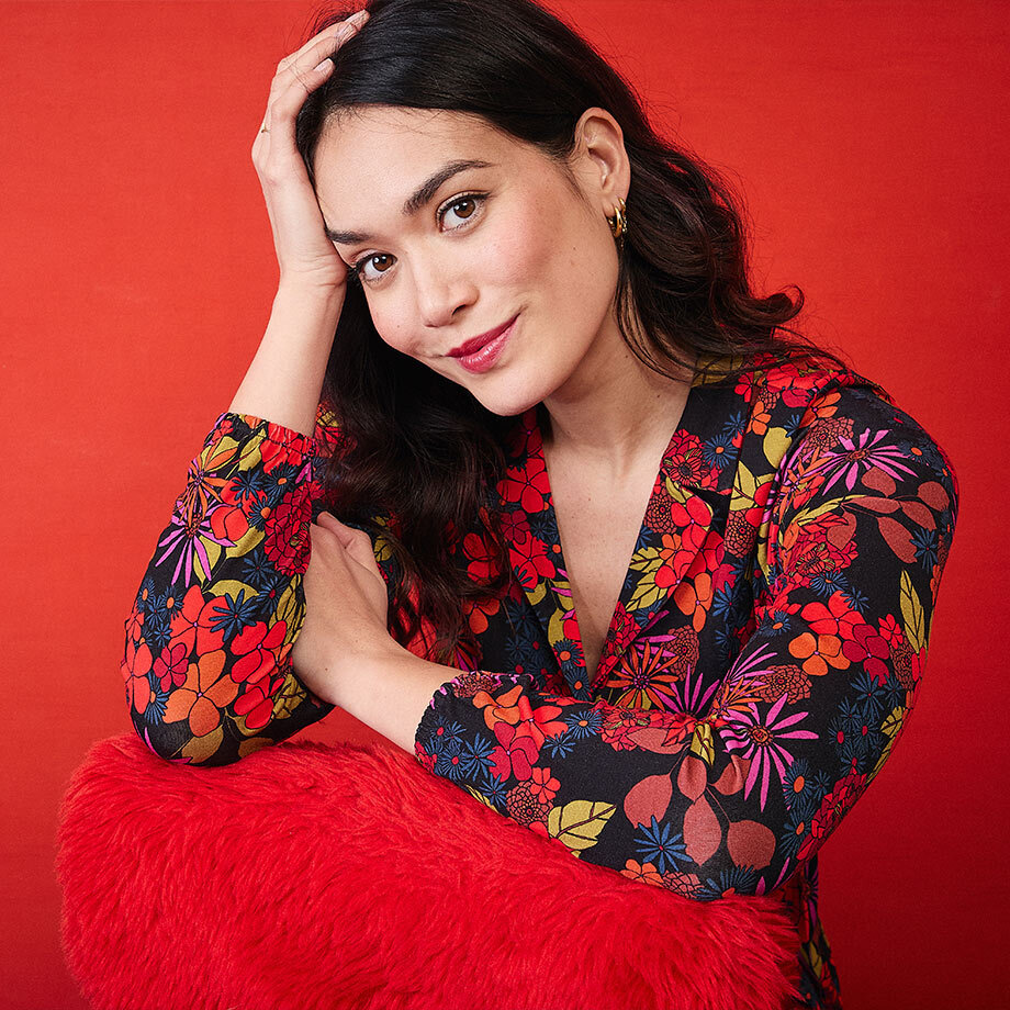 Smiling brunette woman wearing a colorful floral-print dress, leaning on a fluffy red cushion against a vibrant red background.