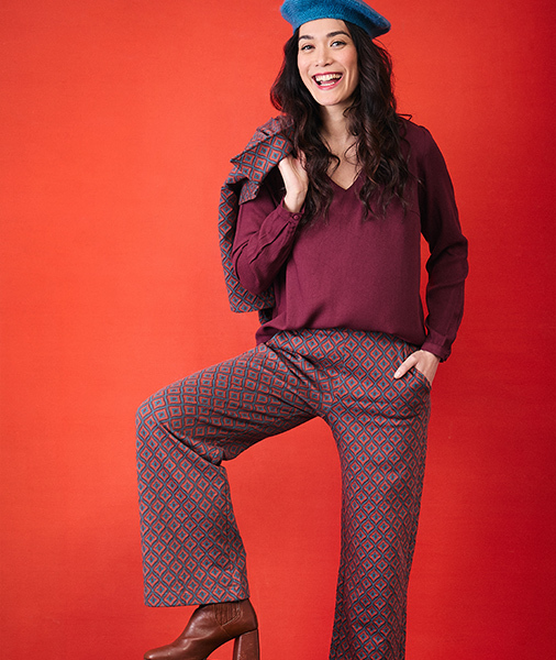 smiling-woman-wearing-a-blue-beret-a-burgundy-blouse-and-blue-and-red-geometric-patterned-trousers-posing-against-a-bright-red-orange-background-