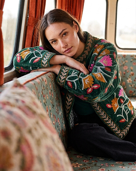 woman-seated-on-a-vintage-style-train-bench-resting-on-her-arms-and-wearing-a-green-jacket-embroidered-with-colorful-floral-motifs-