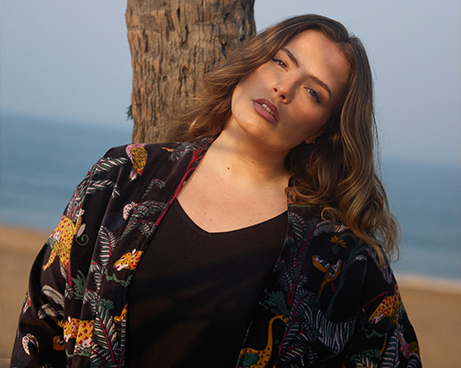 model-with-wavy-brown-hair-leaning-against-a-tree-by-the-sea-wearing-a-black-top-and-a-flowing-black-jacket-with-a-colorful-print-