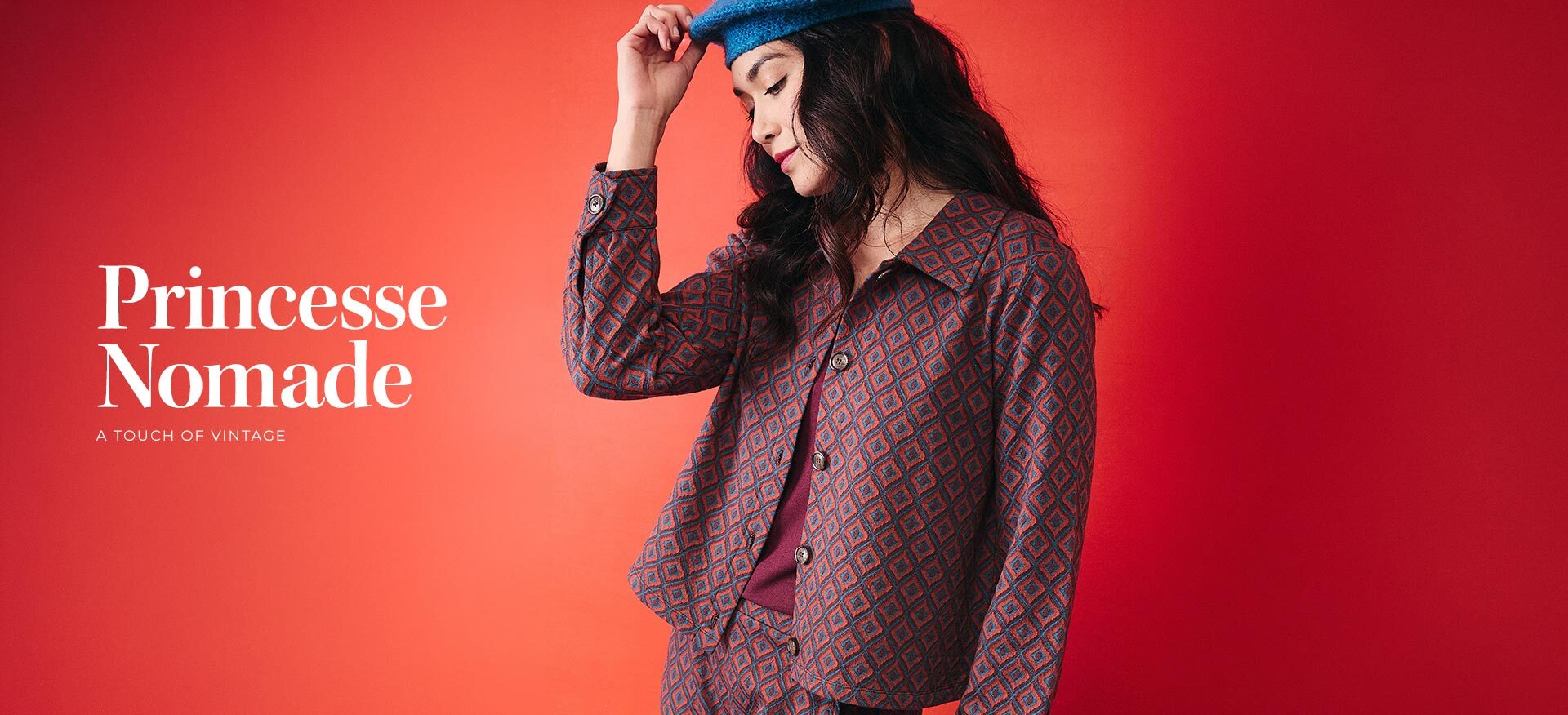 model-wearing-a-red-and-blue-geometric-print-jacket-and-trousers-with-a-blue-beret-against-a-red-orange-gradient-background--princesse-nomade-