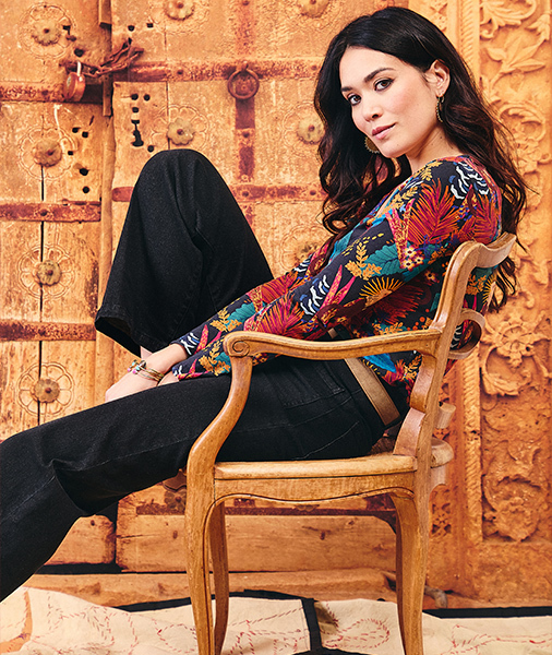 a-woman-with-long-wavy-brown-hair-is-sitting-on-a-carved-wooden-chair-posing-in-front-of-an-ancient-ornate-wooden-door--she-is-wearing-a-long-sleeved-top-with-a-tropical-floral-pattern-and-wide-leg-black-pants--she-is-looking-directly-at-the-camera-with-a-slight-smile-her-arm-resting-on-the-chair-s-armrest-