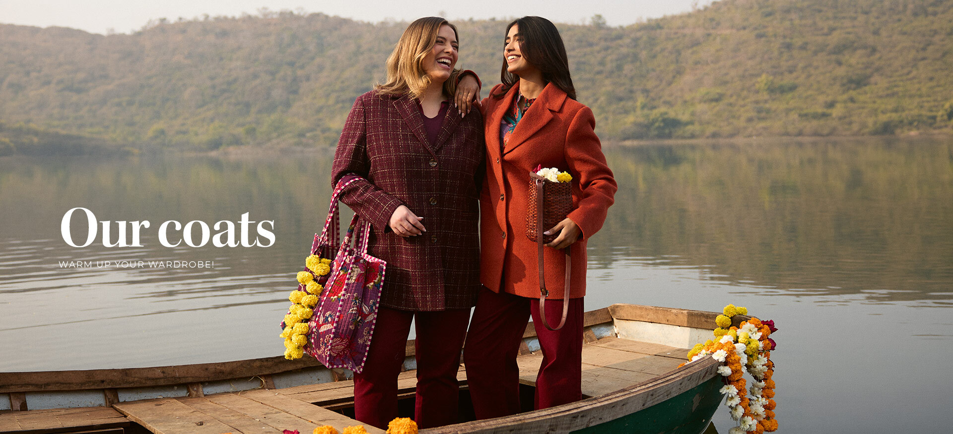 two-smiling-women-wearing-winter-coatsone-in-a-burgundy-plaid-coat-and-the-other-in-a-bright-orange-coatstanding-on-a-wooden-boat-on-a-lake-surrounded-by-hills--text-overlay_--our-coats---warm-up-your-wardrobe--
