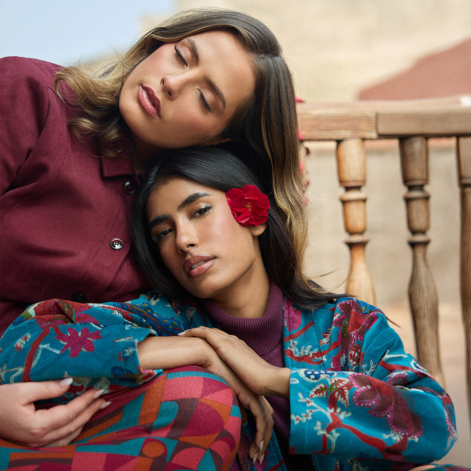 Two smiling women photographed from a low angle, wearing colorful outfits with graphic and floral prints, one holding red flowers in the foreground.