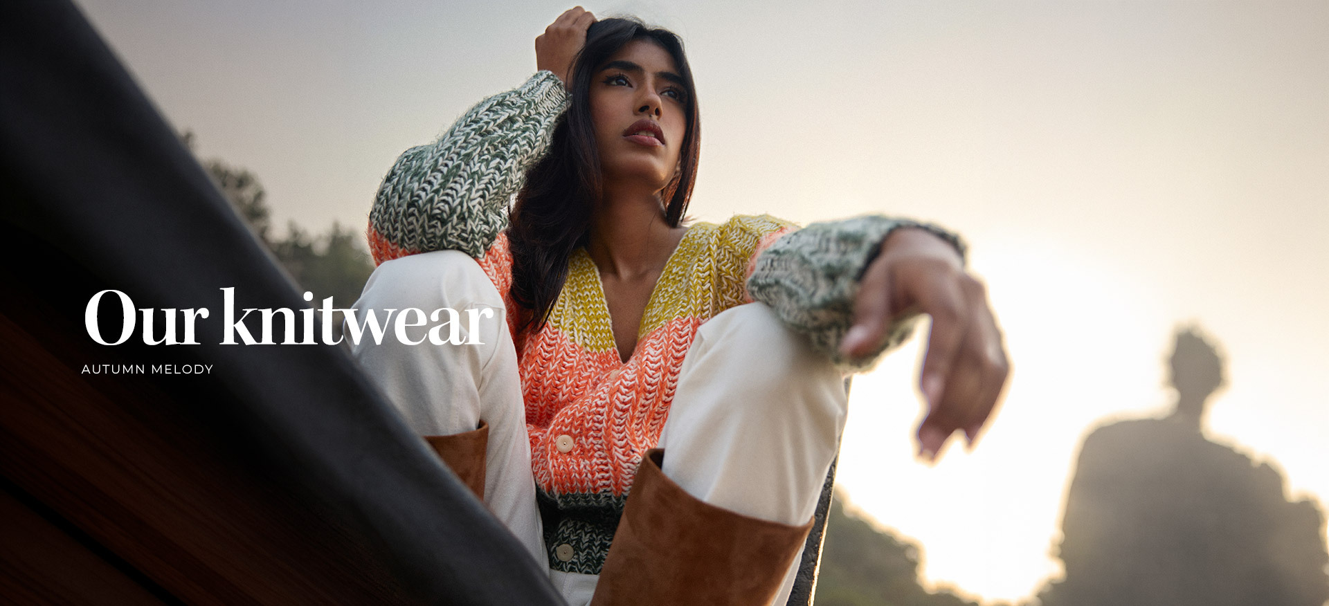 model-sitting-outdoors-at-sunset-wearing-a-chunky-multicolored-knit-cardigan-in-yellow-coral-and-green-paired-with-white-trousers-and-tan-boots--our-knitwear--autumn-melody-collection-